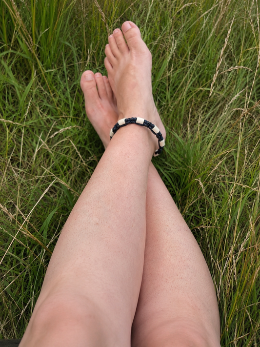 A person's legs and feet resting in green grass, wearing a beaded ankle bracelet with alternating black and white beads.