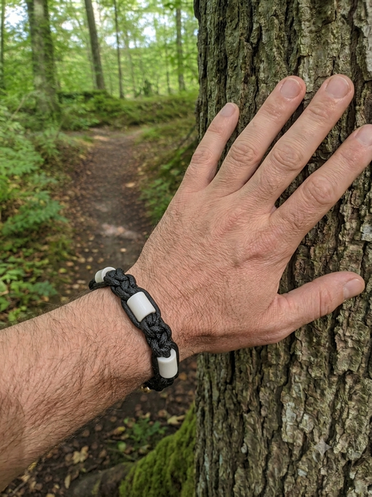 A black braided leather bracelet with white rectangular beads worn on a person's wrist, shown in a forest setting with their hand resting against a tree trunk.