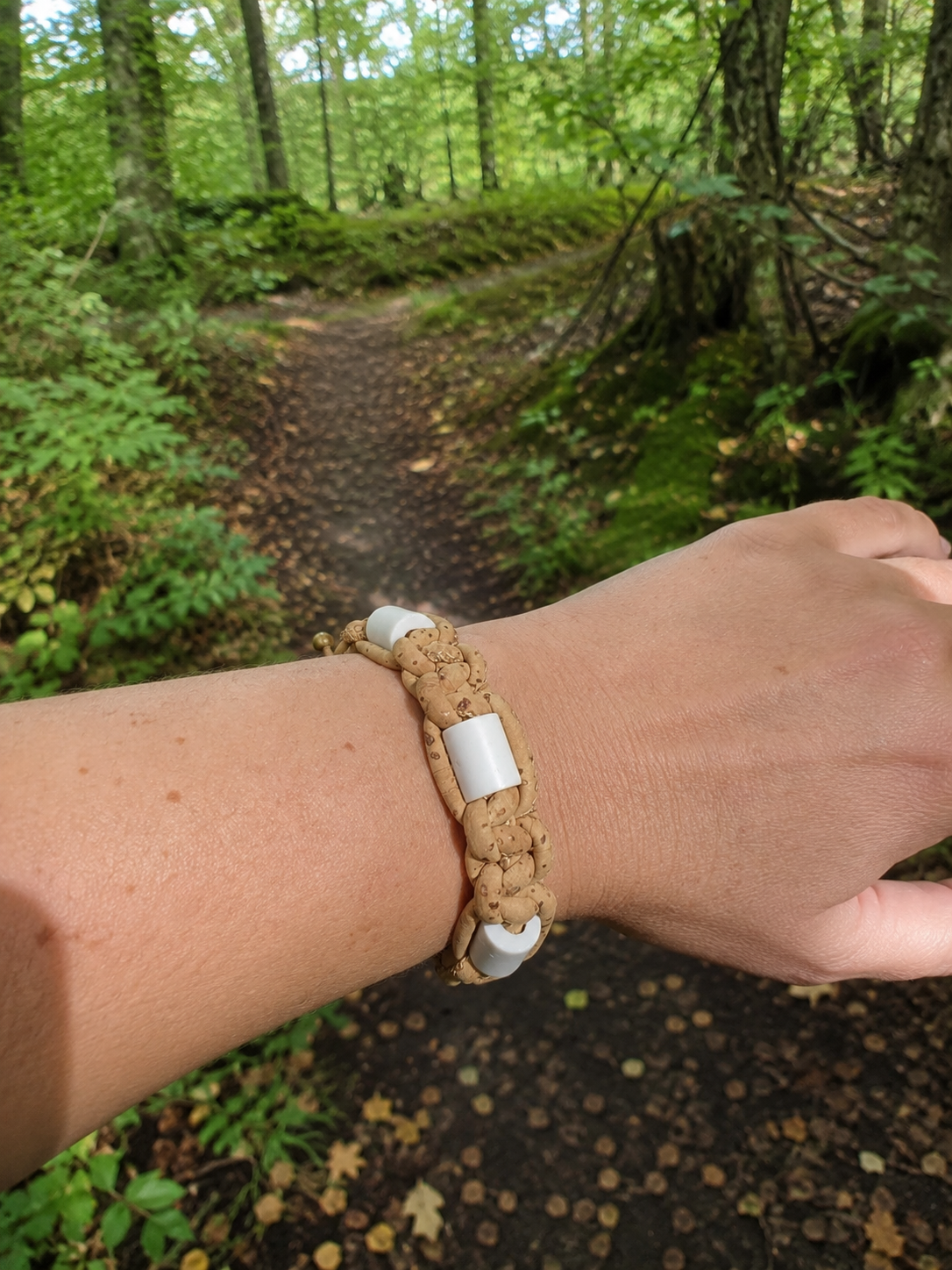 A tan woven bracelet with white rectangular beads worn on a person's wrist, photographed outdoors on a forest path.